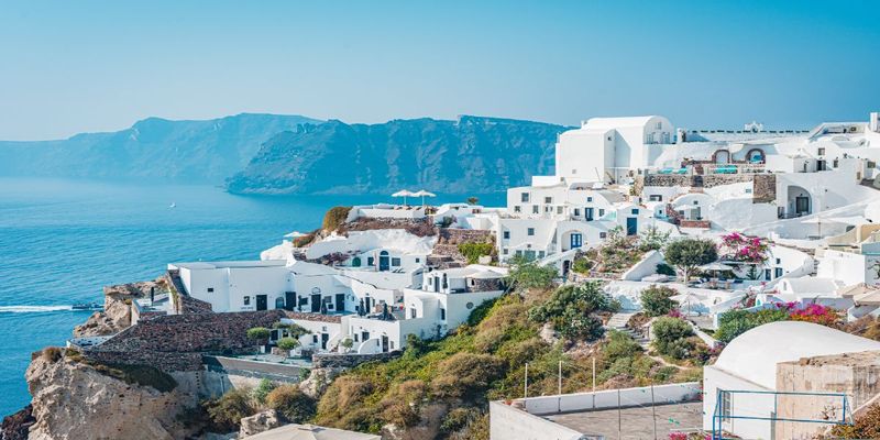White-washed buildings with blue domes on a clifftop overlooking turquoise sea and distant caldera