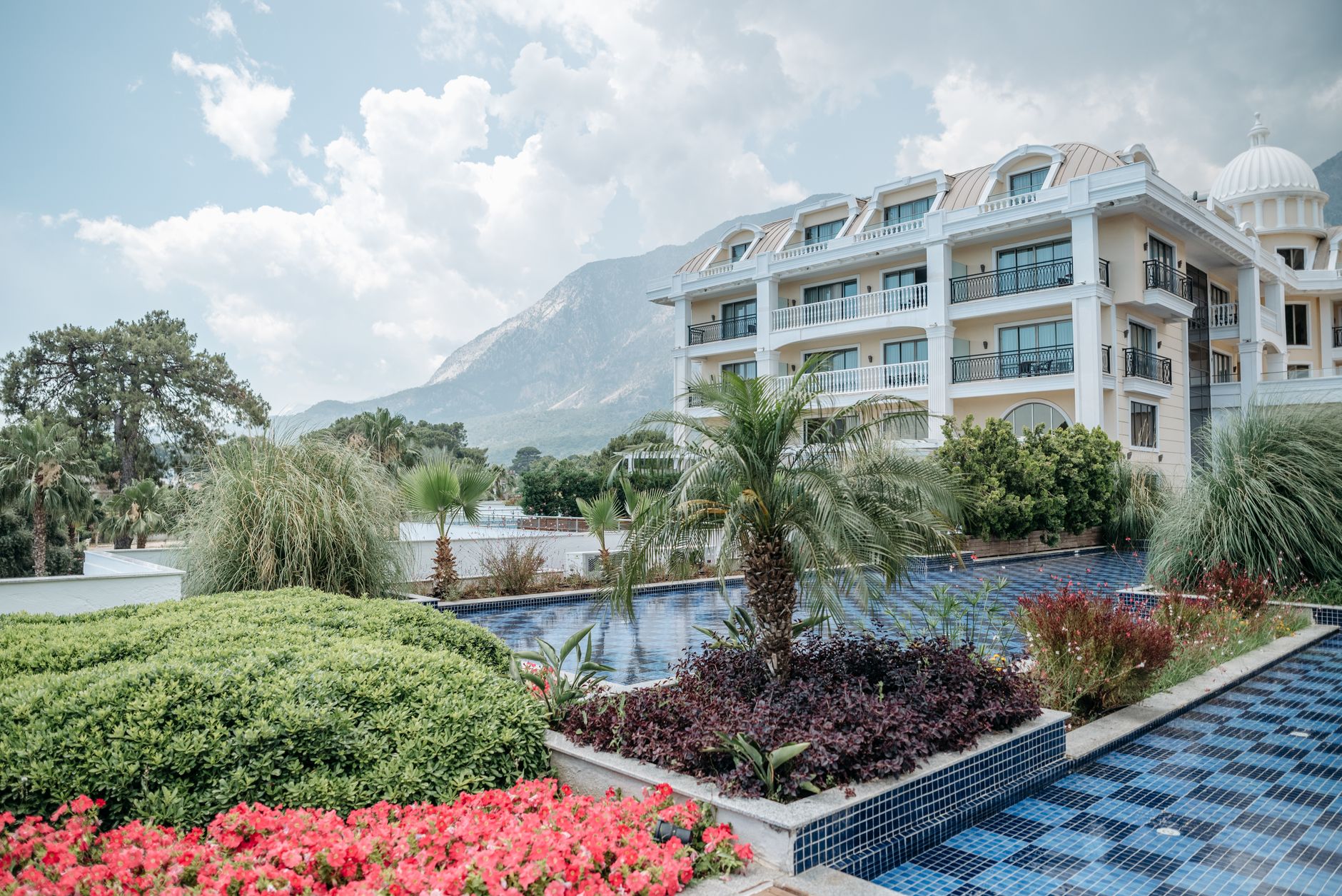 Hôtel blanc avec dôme, piscine bleue et jardins fleuris face à une montagne