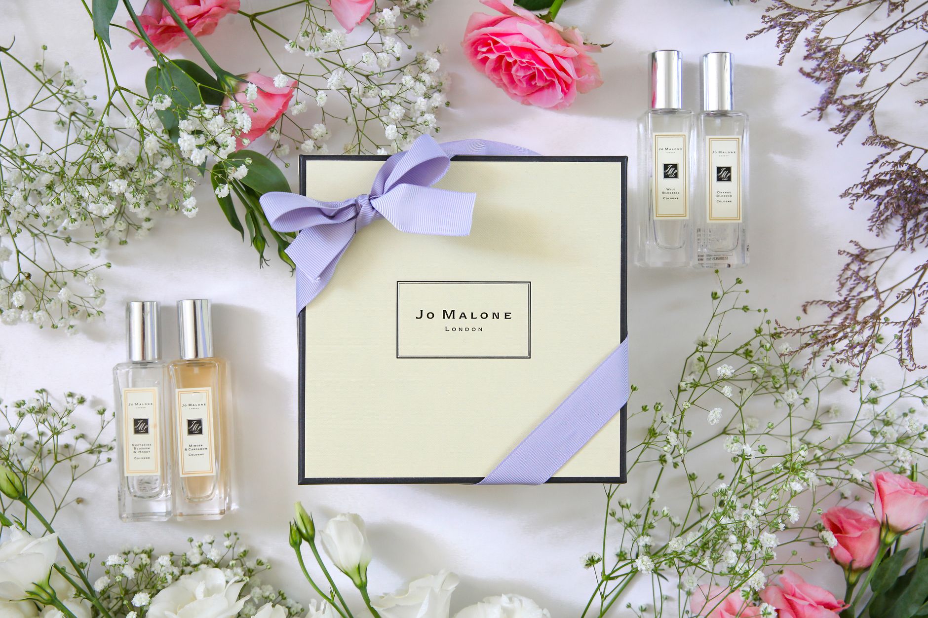 Jo Malone London fragrance gift set with two bottles surrounded by pink roses, baby's breath, dried flowers, and purple ribbon on white background