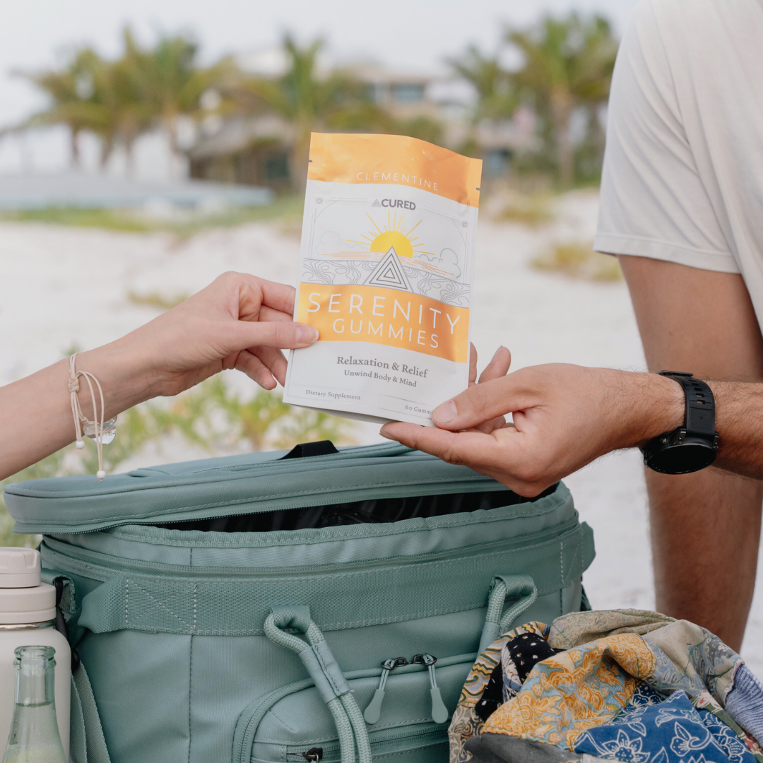 Two people holding a package of Clementine Serenity Gummies on a beach with palm trees and cooler visible