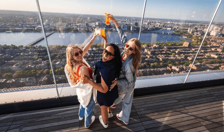 Three women holding colorful drinks while posing at an outdoor observation deck overlooking a city and waterfront