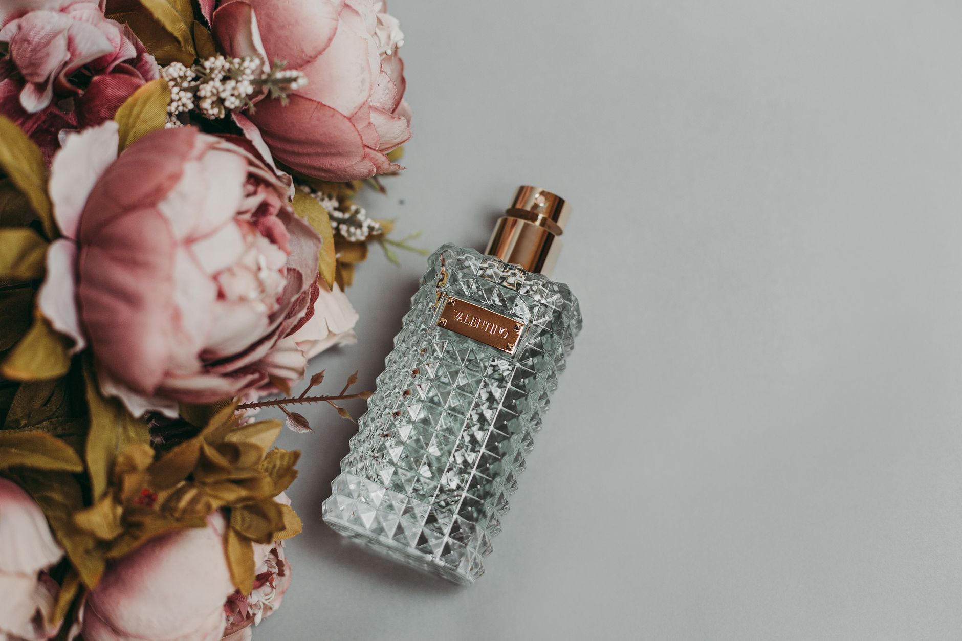 Crystal-cut glass perfume bottle with rose gold atomizer next to pink peony flowers on gray background