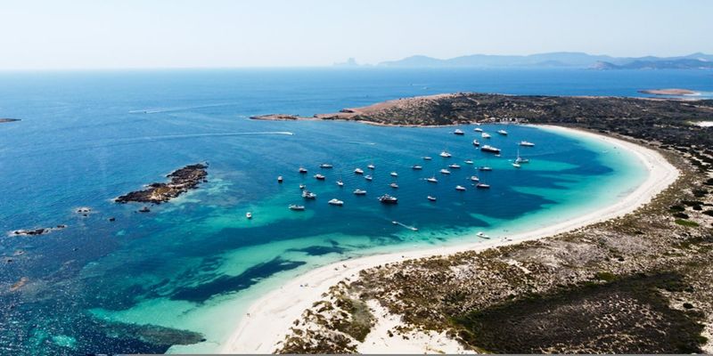 Aerial view of a crescent beach with crystal-clear turquoise water and numerous moored boats