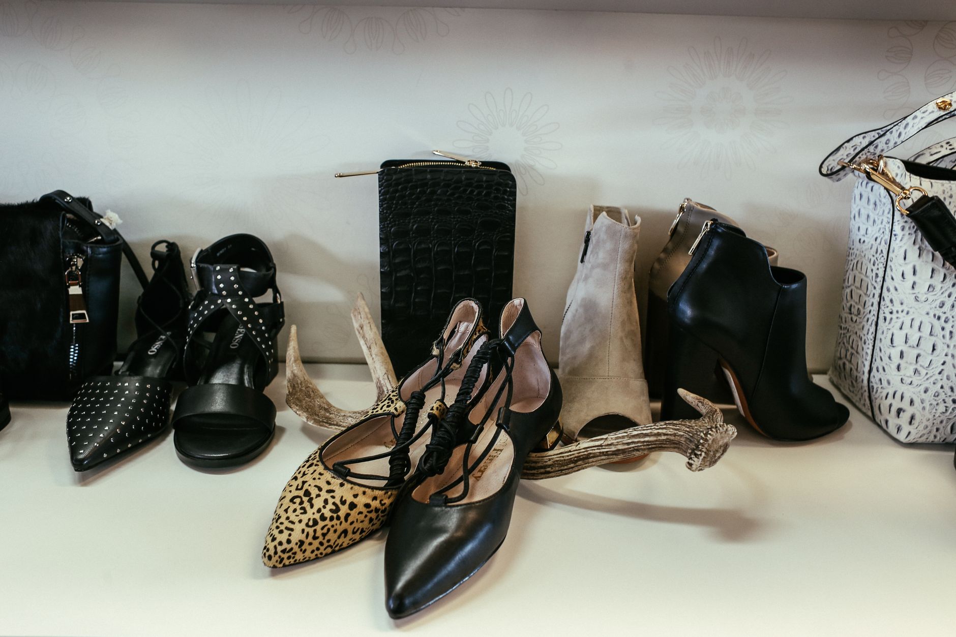 Curated collection of designer shoes and accessories including black studded heels, leopard print flats, crocodile wallet, and patterned clutches arranged on white surface