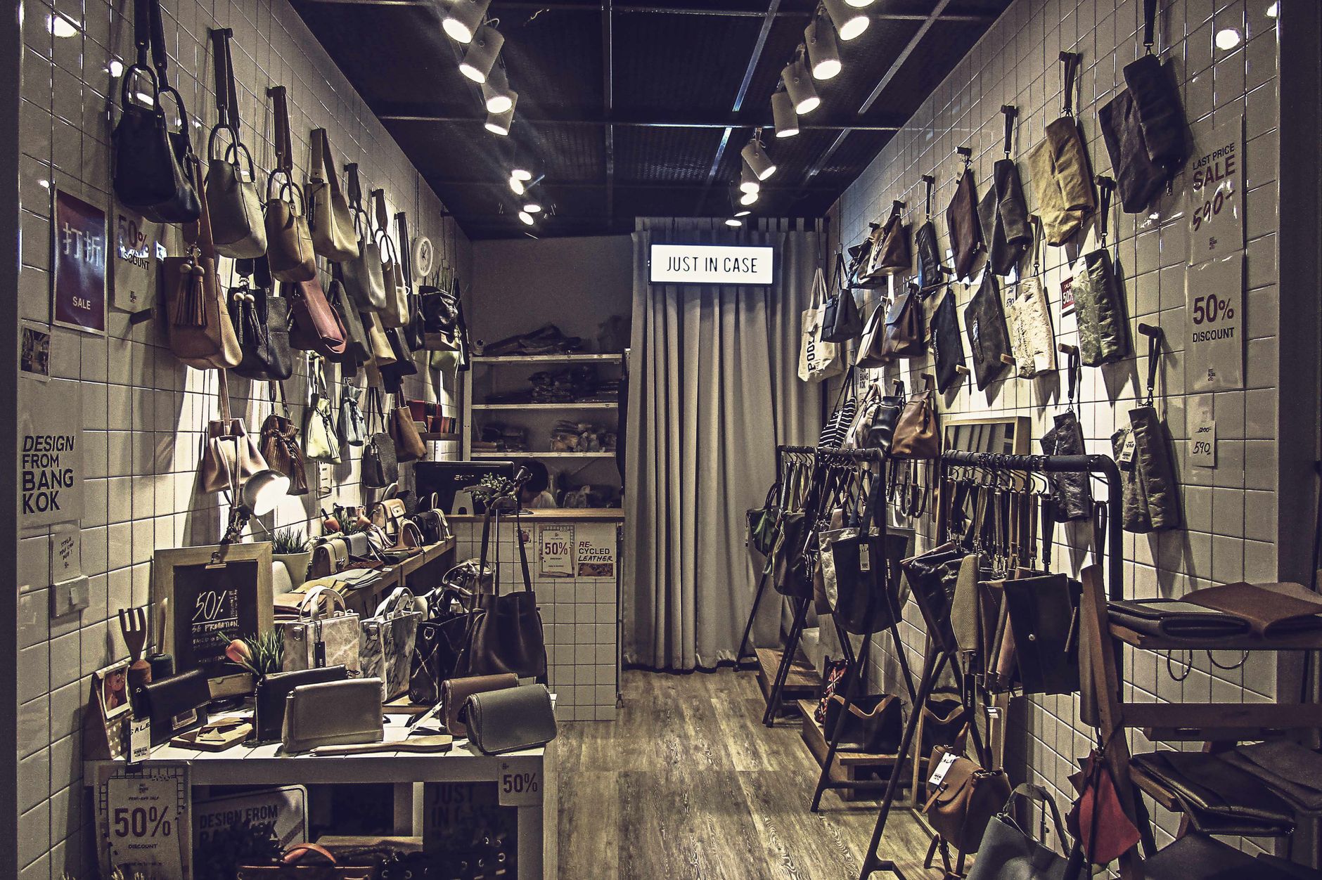 Boutique store interior with white subway tile walls displaying hundreds of hanging leather handbags and accessories with sale signs advertising 50% discounts