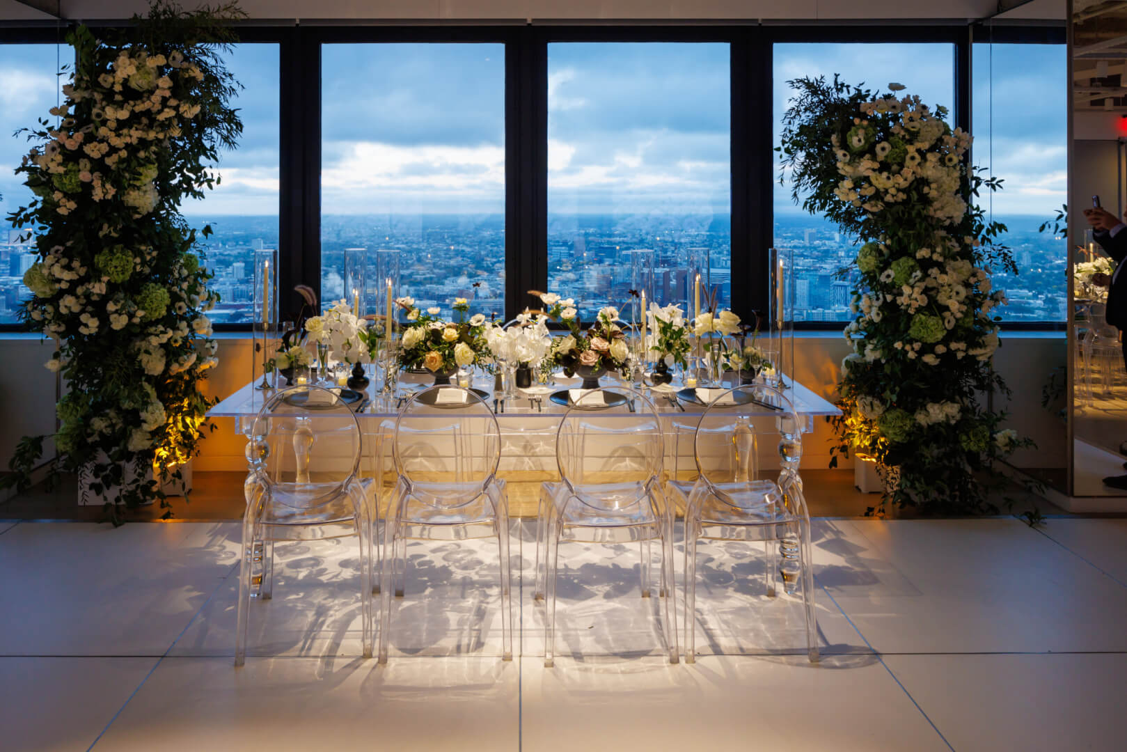 Elegant table setting with floral arrangements overlooking a cityscape through floor-to-ceiling windows