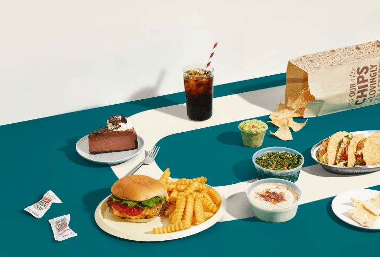 Flat lay of food and drinks on teal and white surface including burger with fries, chocolate cake, tacos, soda, and snack packages