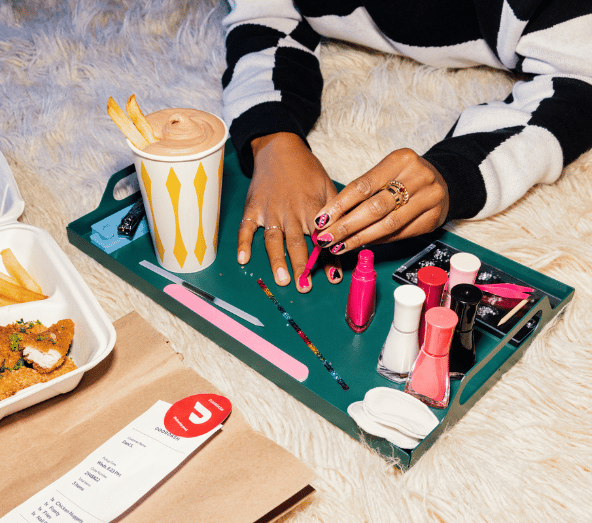 Woman's hands with pink and red nail polish arranging beauty and cosmetic products on teal tray next to beverage and food